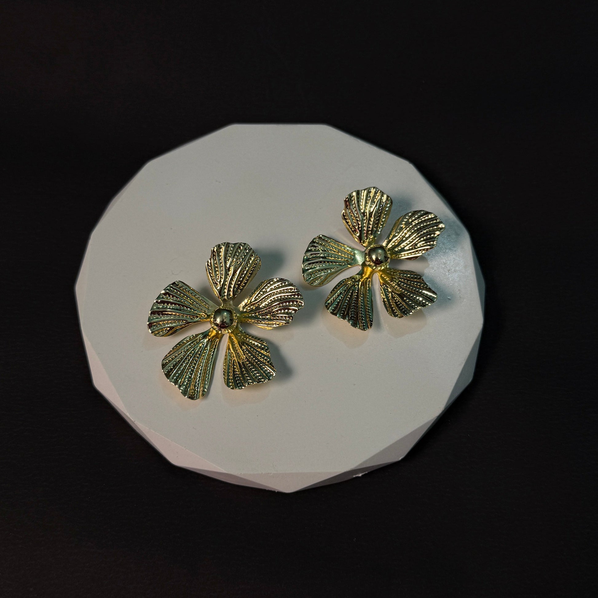 Gold flower-shaped earrings on a white circular stand against a black background